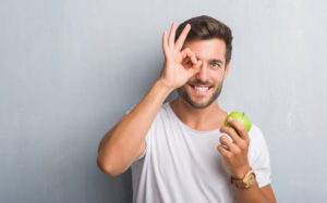 Handsome young man over grey grunge wall eating green apple with happy face smiling doing ok sign with hand on eye looking through fingers