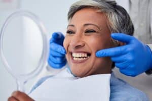Senior woman smiles sitting in the dentist chair, admiring her new crown in the mirror.